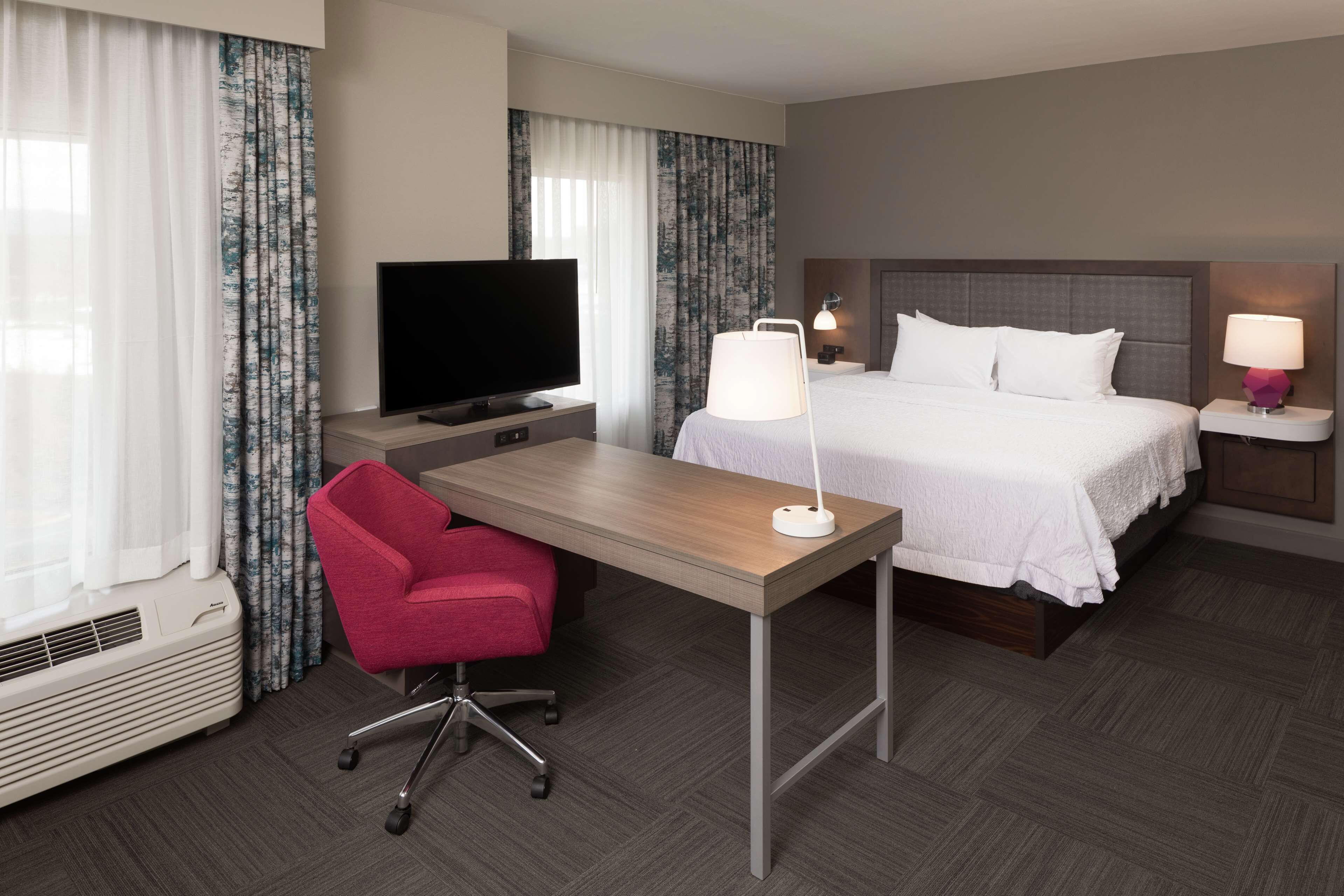 Une chambre d’hôtel comprenant un lit, un bureau avec une chaise, une télévision, deux lampes de chevet et une fenêtre avec des rideaux.