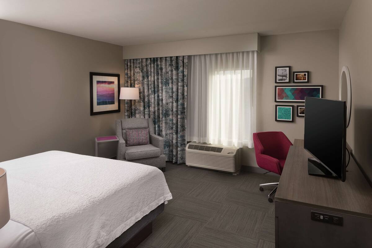 Hotelkamer met een bed, bureau met computer, stoel, raam met gordijnen en een wandairconditioner.