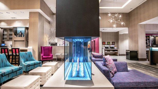 Een moderne hotel lobby met een centrale glazen lift, comfortabele zitplaatsen, een receptiebalie en een bar.