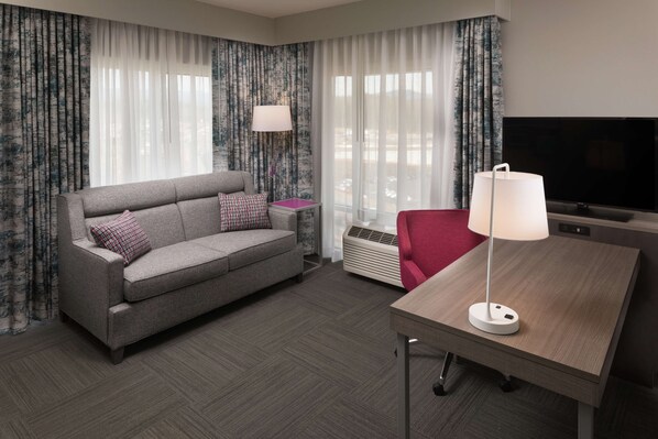 Une chambre d’hôtel avec un canapé gris, un fauteuil rouge, une table en bois, une télévision et une fenêtre avec des rideaux.