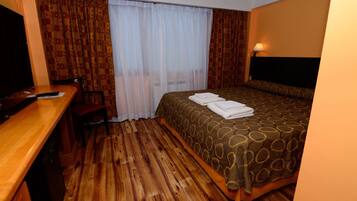 A hotel room with a wooden floor, a bed with a patterned bedspread, a desk with a chair, a television, and a window with curtains.