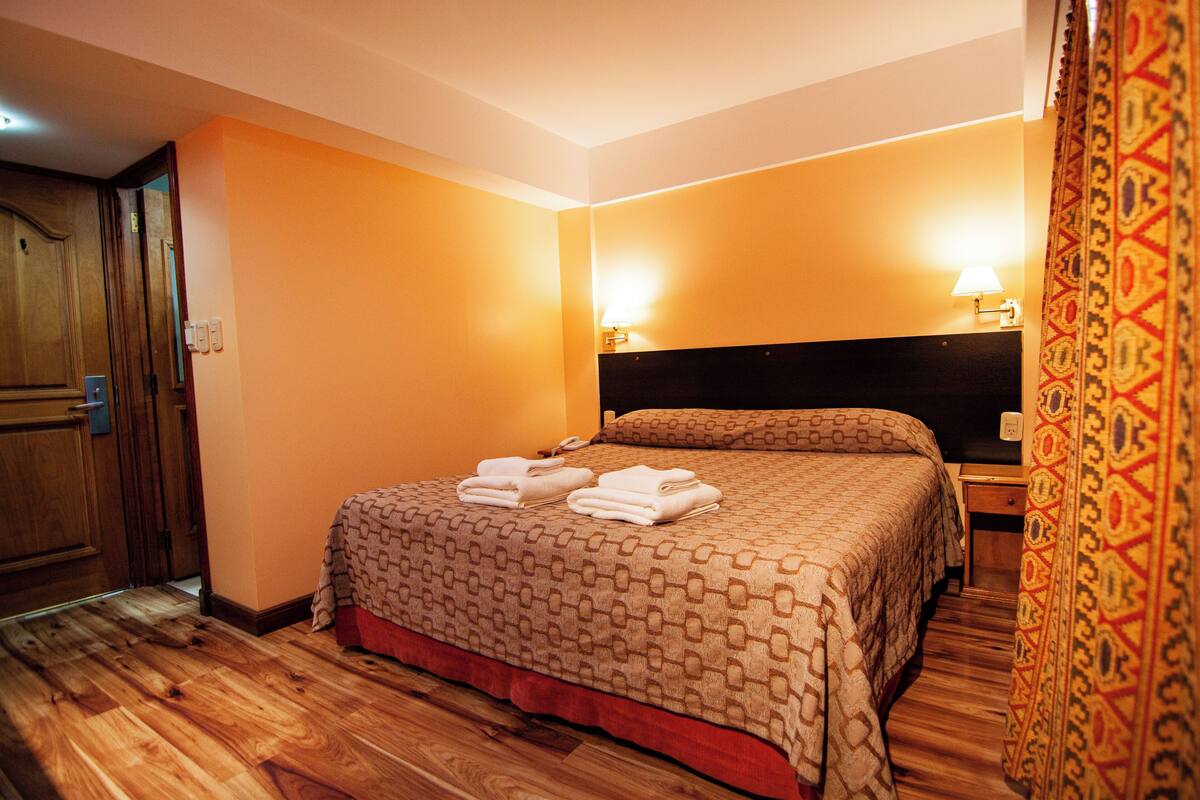 Een hotelkamer met een houten vloer, een bed met een bedsprei met dessin, twee nachtlampjes en een deur.