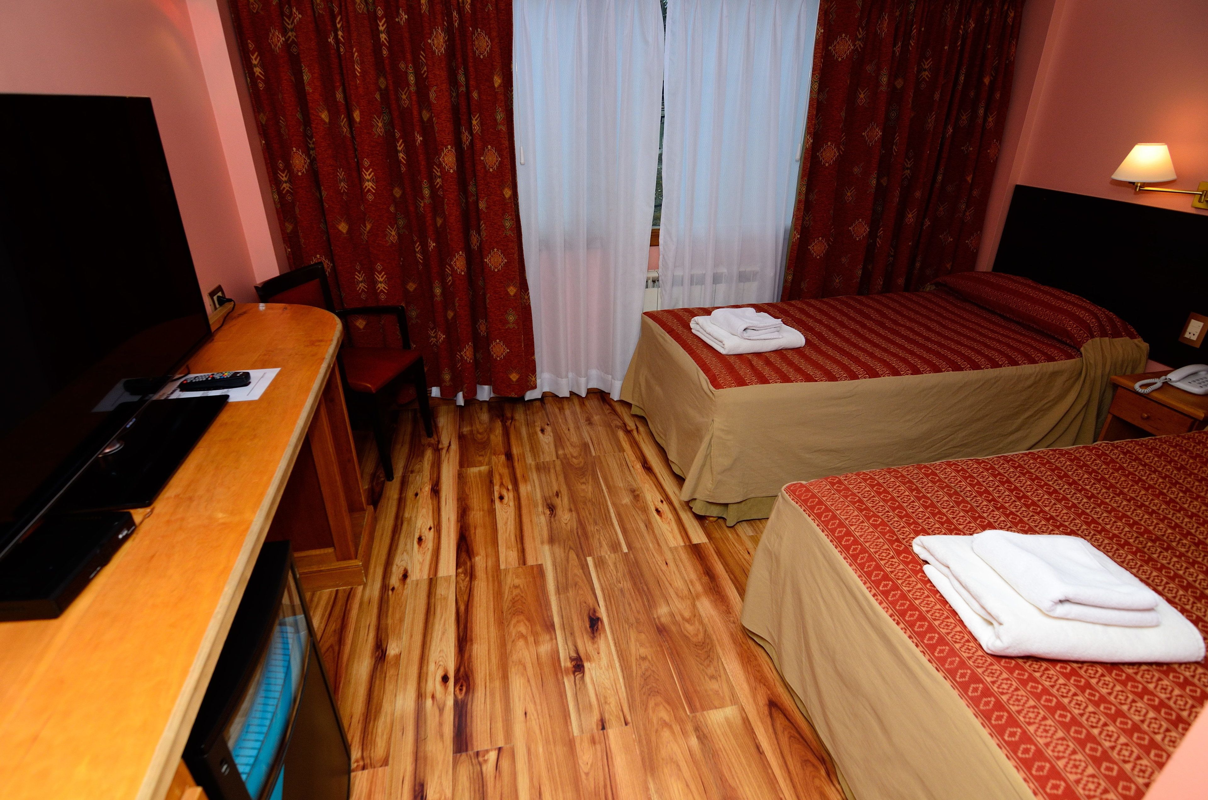 Habitación de hotel con piso de madera, un escritorio con televisor, cama con colcha roja y ventana con cortinas rojas.