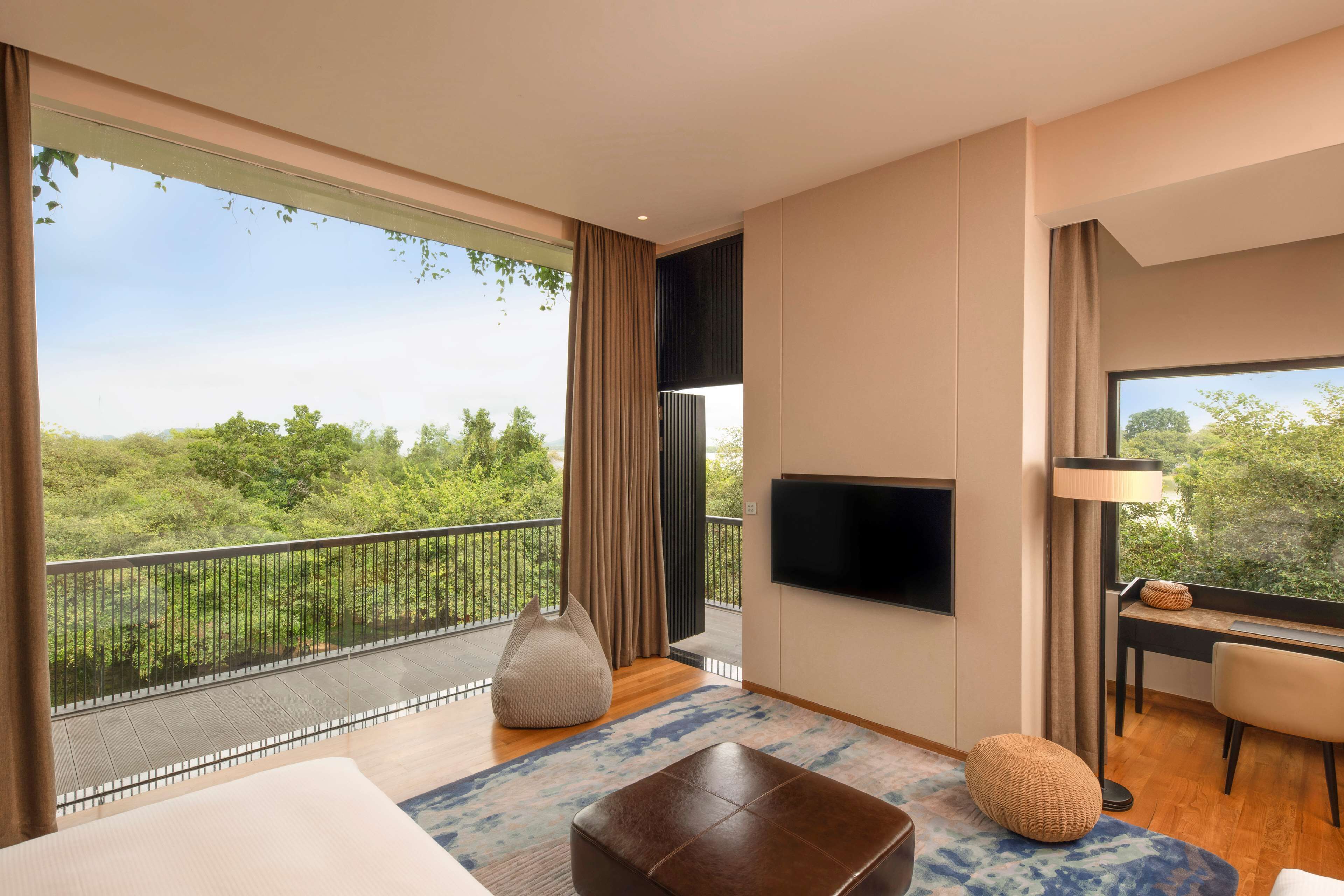 A modern hotel room with a balcony, a flat-screen TV, a desk with a chair, and a large window offering a view of greenery.