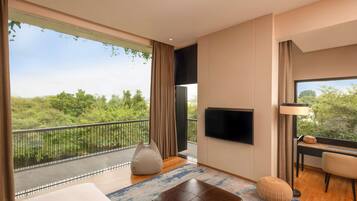 A modern hotel room with a balcony, a flat-screen TV, a desk with a chair, and a large window offering a view of greenery.