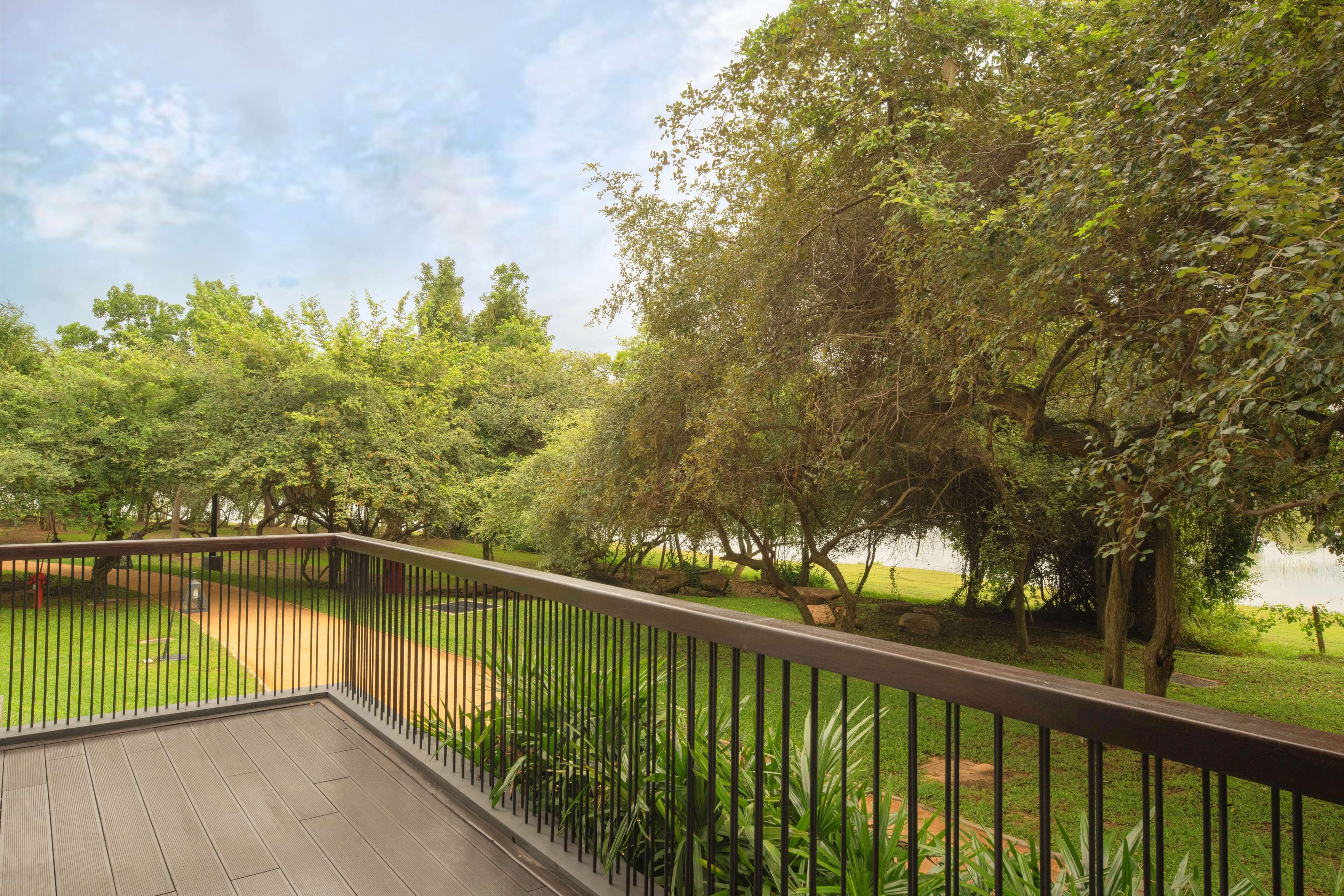 Une terrasse en bois avec une rambarde en métal, donnant sur une zone herbeuse parsemée d’arbres et bordée d’un plan d’eau.