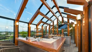 A modern bar area with a wooden counter, bar stools, and a glass enclosure with a view of the outdoors. - DoubleTree by Hilton Weerawila Rajawarna Resort (Weerawila)