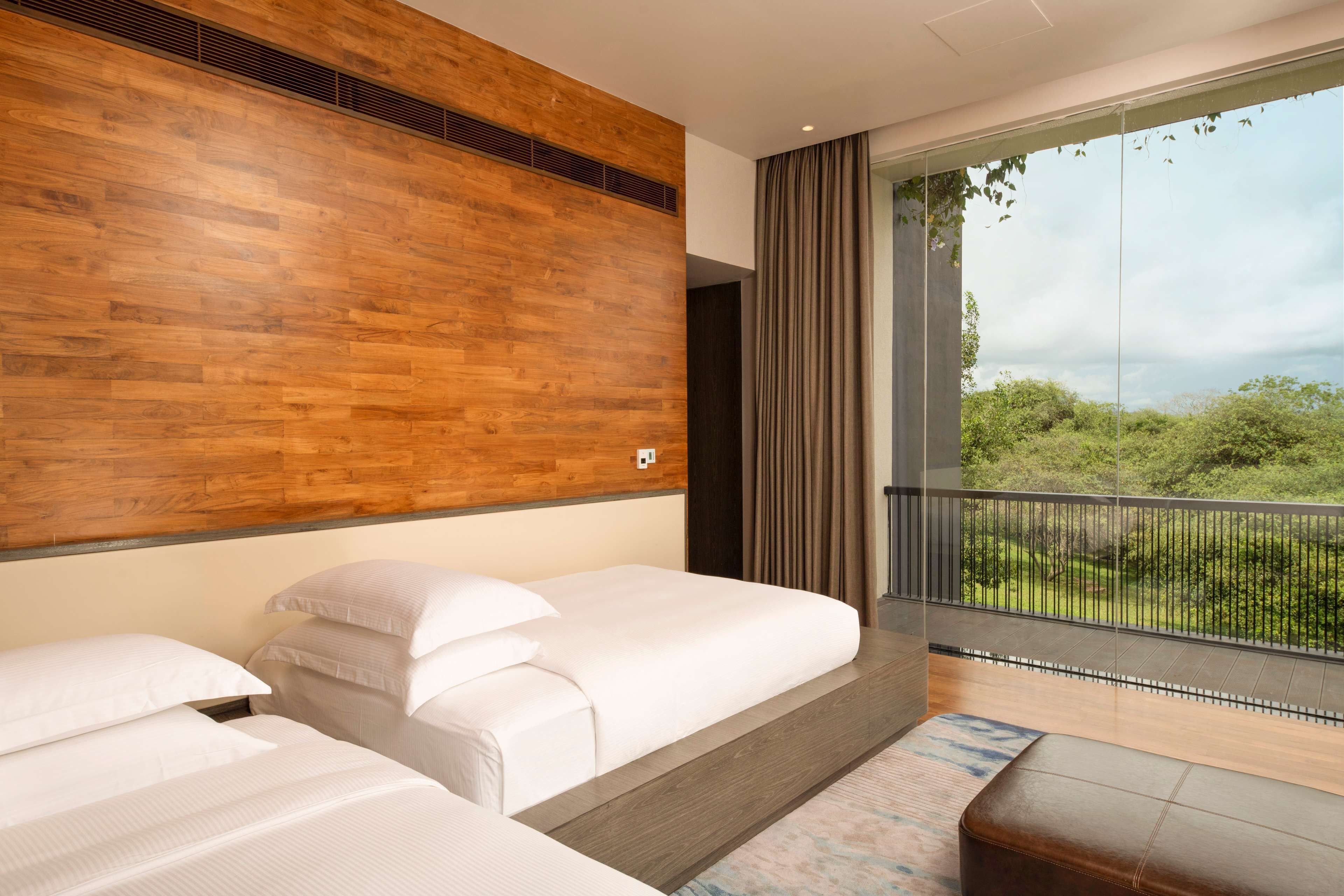 A hotel room with two beds, a wooden accent wall, a balcony with a view of greenery, and a modern design.
