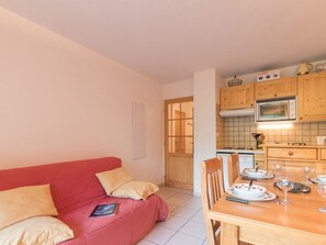 Une pièce chaleureuse avec un canapé rouge, une table à manger en bois mise avec des assiettes et des verres, et un coin cuisine doté de meubles en bois et d’un four à micro-ondes.