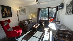 A living room with a red chair, a brown sofa, and a dining table with chairs. There are two windows with curtains, a ceiling light, and a wall-mounted TV. - Cozy studio with mountain nook for 4 people in Chantemerle (Saint-Chaffrey)