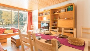 Une pièce chaleureuse et bien éclairée, avec une table à manger en bois préparée pour deux personnes, entourée d’étagères à livres et d’un téléviseur.