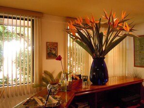 A room with a wooden table, a vase with a red rose, and a large plant with orange leaves. - Hostal Verona (San Salvador)