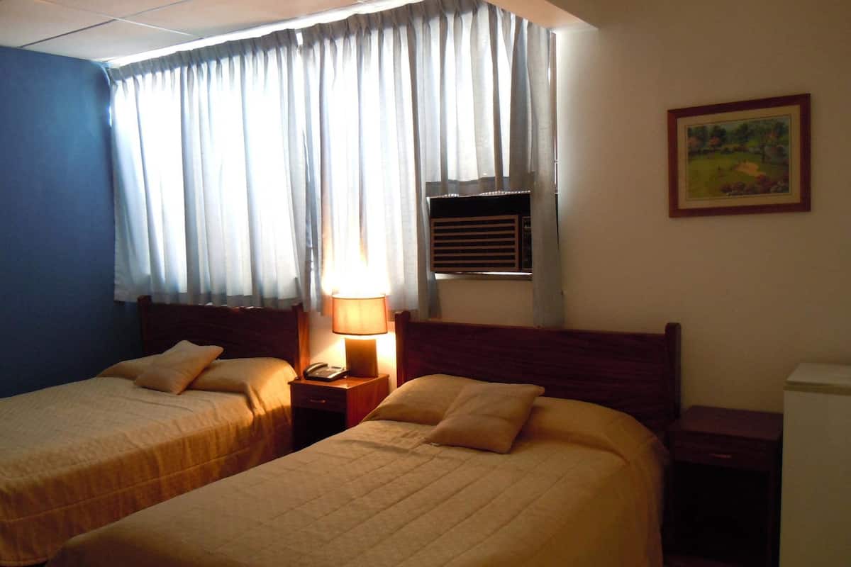 A hotel room with two beds, a nightstand with a lamp, a window with curtains, and a painting on the wall.