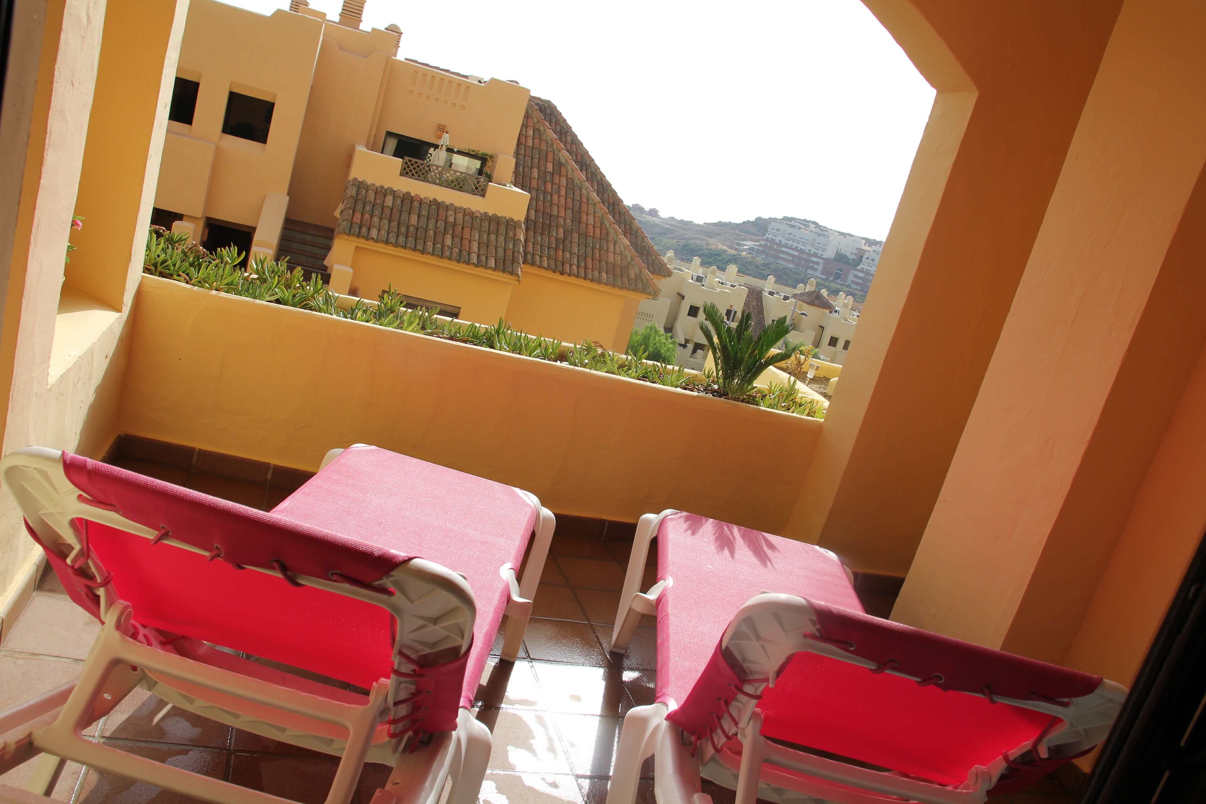 A balcony with two red chairs and a table, overlooking a residential area with buildings and a hill in the background.