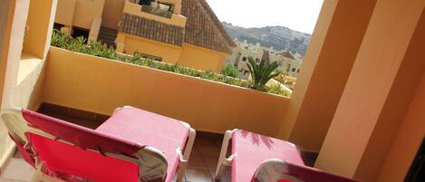 A balcony with two red chairs and a table, overlooking a residential area with buildings and a hill in the background.
