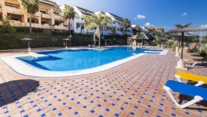 A swimming pool with a tiled deck, lounge chairs, palm trees, and a building with balconies. - Apartment cozy in quiet area near of beaches and shops (Manilva)
