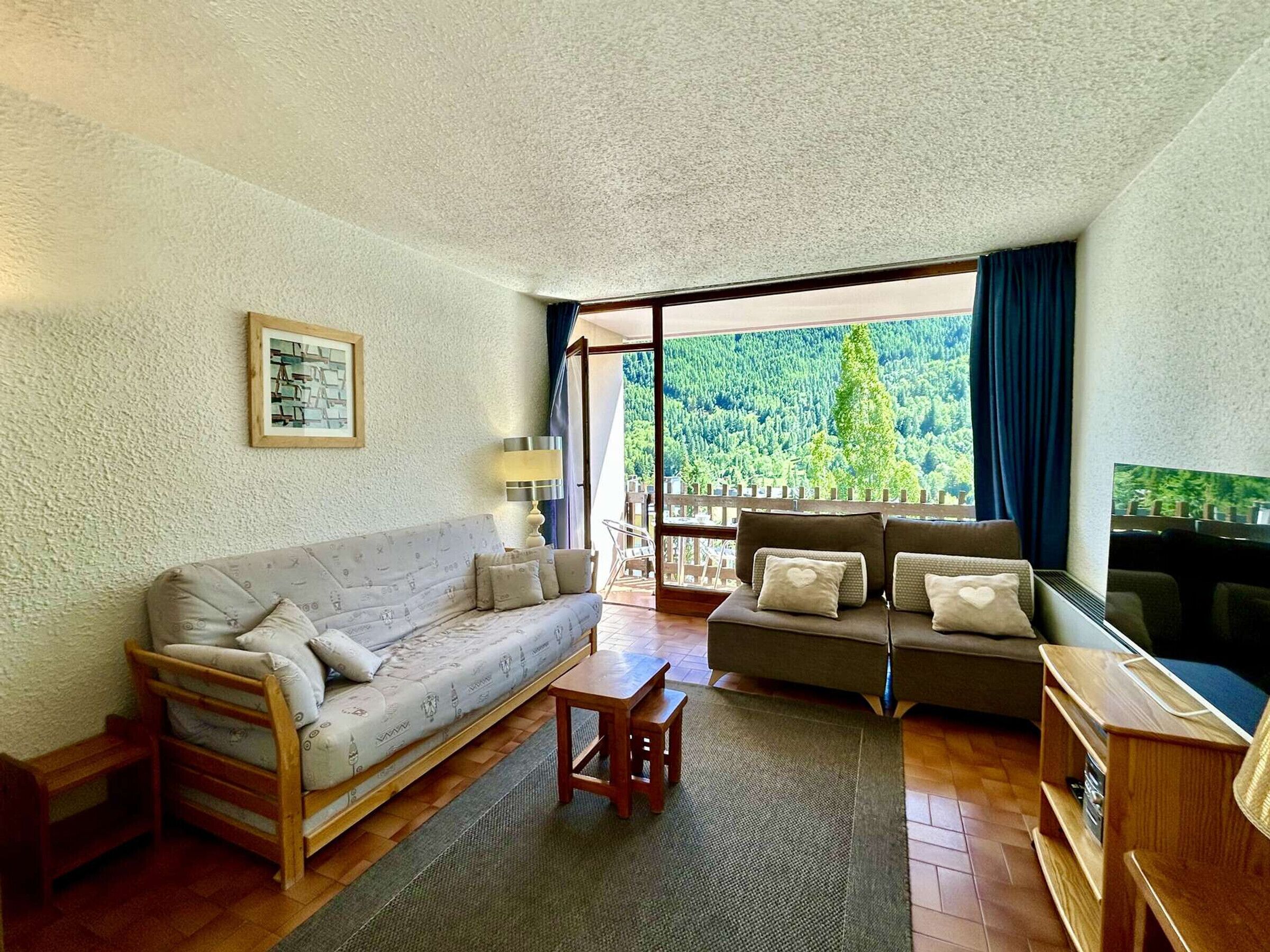 A cozy living room with a sofa, a coffee table, and a TV. There is a large window with a view of trees and a balcony.