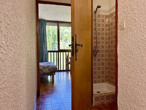 A room with a wooden door leading to a balcony with a bed and a view of greenery.