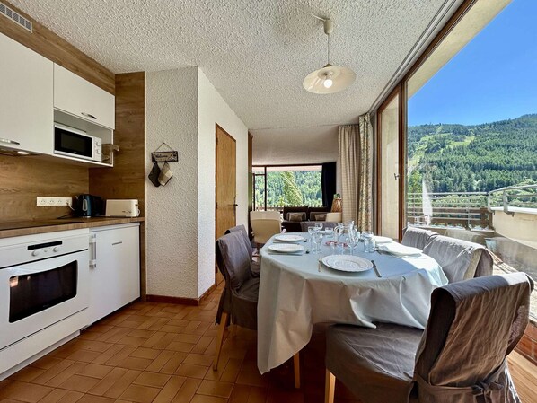 Un coin repas avec une table dressée pour quatre personnes, une cuisine équipée d’appareils électroménagers blancs et une vue panoramique sur une colline boisée.