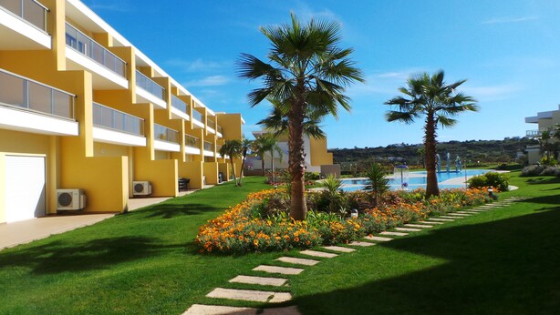 Un hotel con fachada amarilla, piscina y jardín bien cuidado con palmeras y flores.