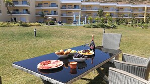 Mesa de comedor al aire libre con comida y bebidas, rodeada de sillas de mimbre, frente a un hotel con balcones y piscina.