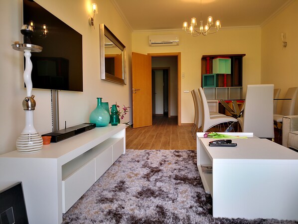 Un salon moderne avec un meuble de rangement blanc pour les appareils électroniques, un grand miroir, un lustre et une vue sur la salle à manger.