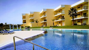 Una piscina con plataforma de baldosas, sillas de descanso y un edificio con balcones al fondo.