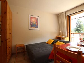 Quarto com cama, uma pequena mesa de madeira com um prato e um copo, uma cadeira, um guarda-roupa de madeira e um quadro na parede.