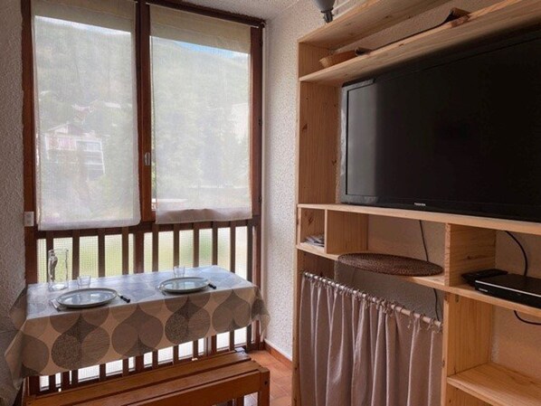 A room with a wooden bench, a table with a patterned tablecloth, a television on a wooden shelf, and a window with a view of greenery.