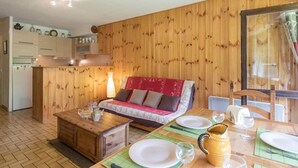 A cozy room with a wooden wall, a sofa, a dining table set for two, and a kitchen area in the background. - Charming 3-room apt for 6 with parking, near slopes and center (Le Monêtier-les-Bains)