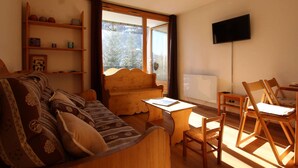 A cozy living room with a sofa, wooden coffee table, and chairs. There is a television mounted on the wall and a window with curtains.