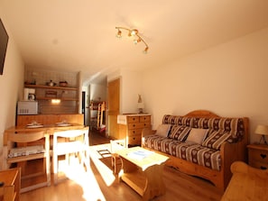 Una habitación acogedora y bien iluminada con una mesa de madera, sillas, un sofá estampado y una kitchenette al fondo.