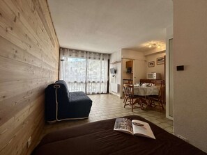 A room with a wooden wall, a sofa, a dining table with chairs, and a kitchen area.