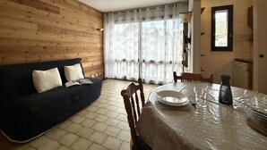 Ett rum med en väggpanel av trä, en mörk soffa, ett matbord med en glasskiva och ett fönster med tunna, genomskinliga gardiner.