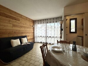 A room with a wooden wall, a dark sofa, a dining table with a glass top, and a window with sheer curtains.