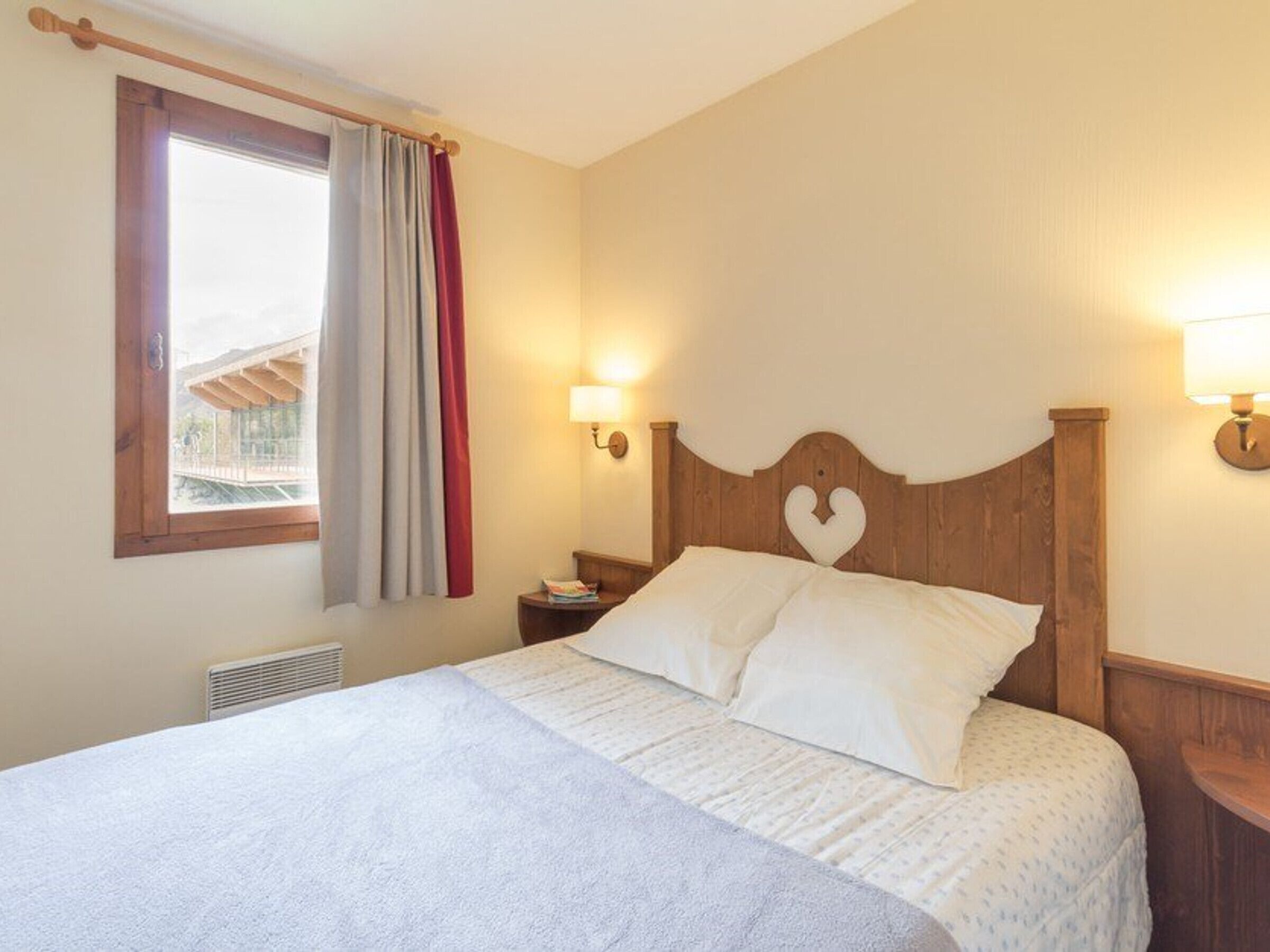 A bedroom with a bed, a window with curtains, a wall-mounted light, and a heater.