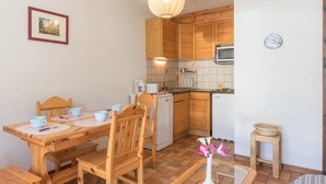 A compact kitchen with wooden cabinets, a dining table with chairs, a microwave, and a small refrigerator. - Cozy 2-Room Apartment with Mountain Views, Balcony & Close to Ski Lifts in Le Monêtier-les-Bains (Le Monêtier-les-Bains)