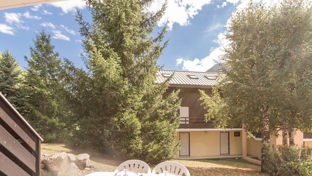 An outdoor dining area with a table set for two, surrounded by trees and a building with a sloped roof. - Charming Studio with Garden, Sleeps 4, South-Facing in Serre-Chevalier (Saint-Chaffrey)
