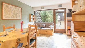A cozy room with a dining table, chairs, a sofa, a bookshelf, and a large window offering a view of greenery outside. - Charming Studio with Garden, Sleeps 4, South-Facing in Serre-Chevalier (Saint-Chaffrey)