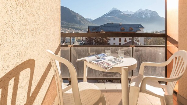 A balcony with two chairs and a table, overlooking a mountain range and a town. - Cozy 2-Room Apartment for 4-6, Near Prorel Cable Car, South-Facing Balcony, Ski Locker (Briançon)