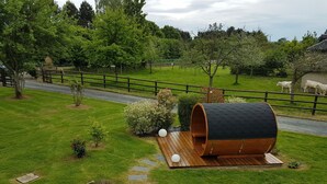 A rustic wooden barrel structure with a wooden deck, surrounded by a fenced area with a gravel path, and a grassy field with trees and a fence in the background.