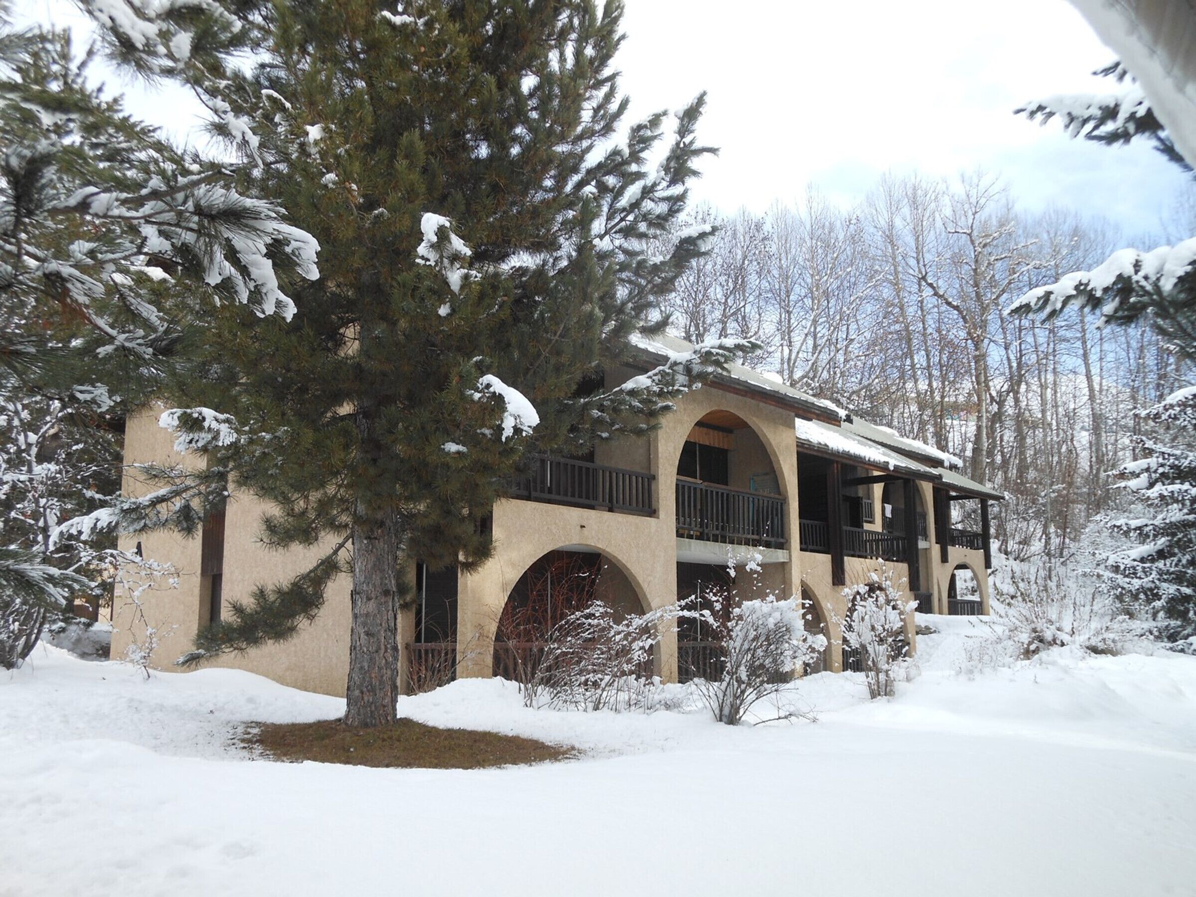 Um edifício coberto de neve com varanda e janelas em arco, rodeado por árvores e neve.