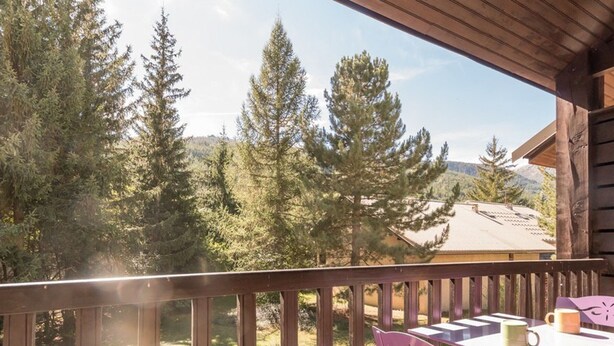 Una terraza de madera con una mesa y sillas, con vista a una zona boscosa de árboles altos y un edificio al fondo.