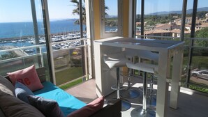 Un balcon avec vue sur la mer et le port de plaisance, une petite table avec des tabourets de bar et un canapé avec des coussins.