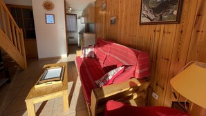 A cozy room with a wooden interior, a red sofa with patterned cushions, a wooden table, and a kitchen area in the background. - Duplex for 6 in Serre-Chevalier - Chantemerle (Saint-Chaffrey)
