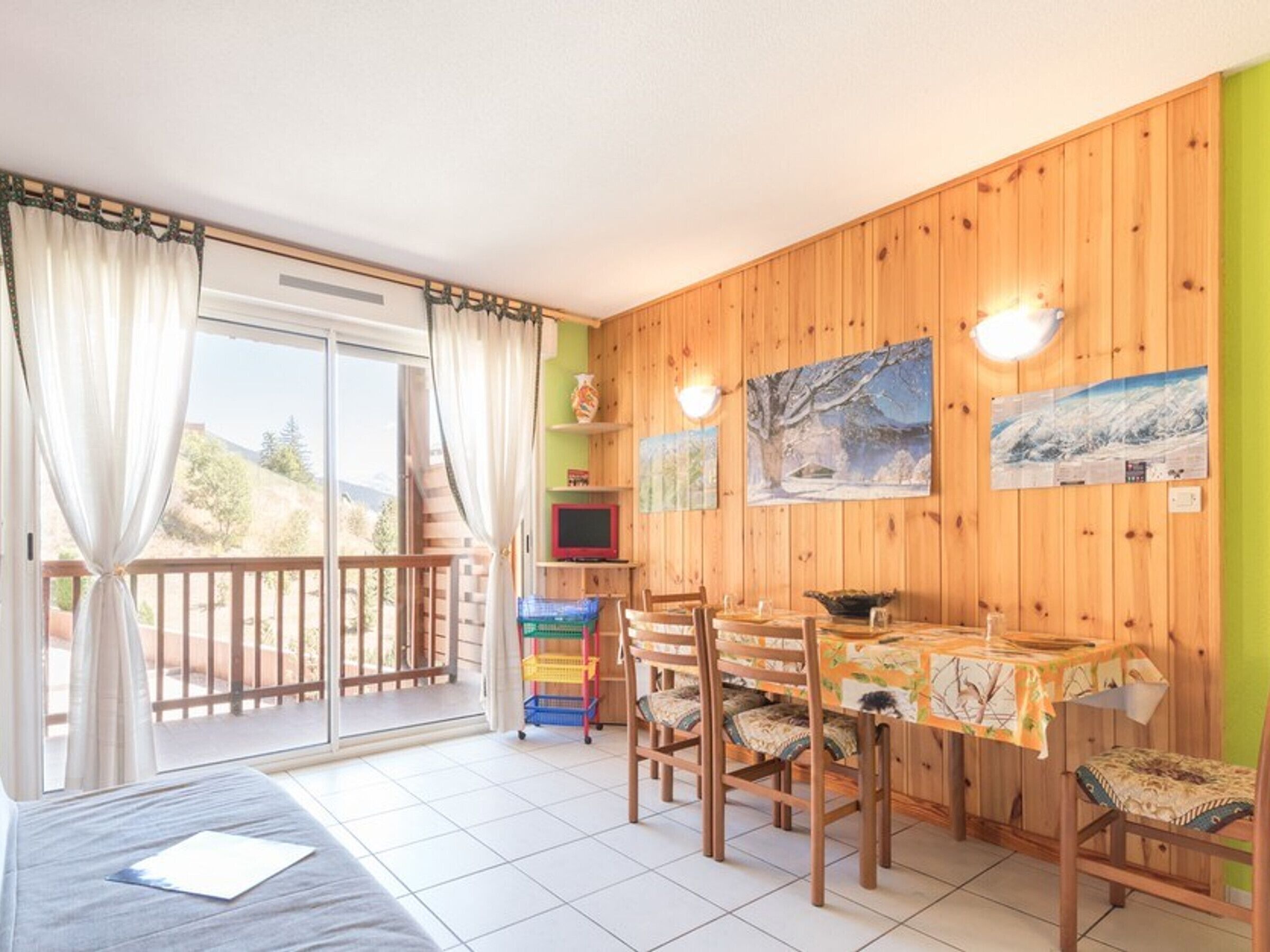 A room with a dining table, chairs, a bed, a TV, and a balcony with curtains.