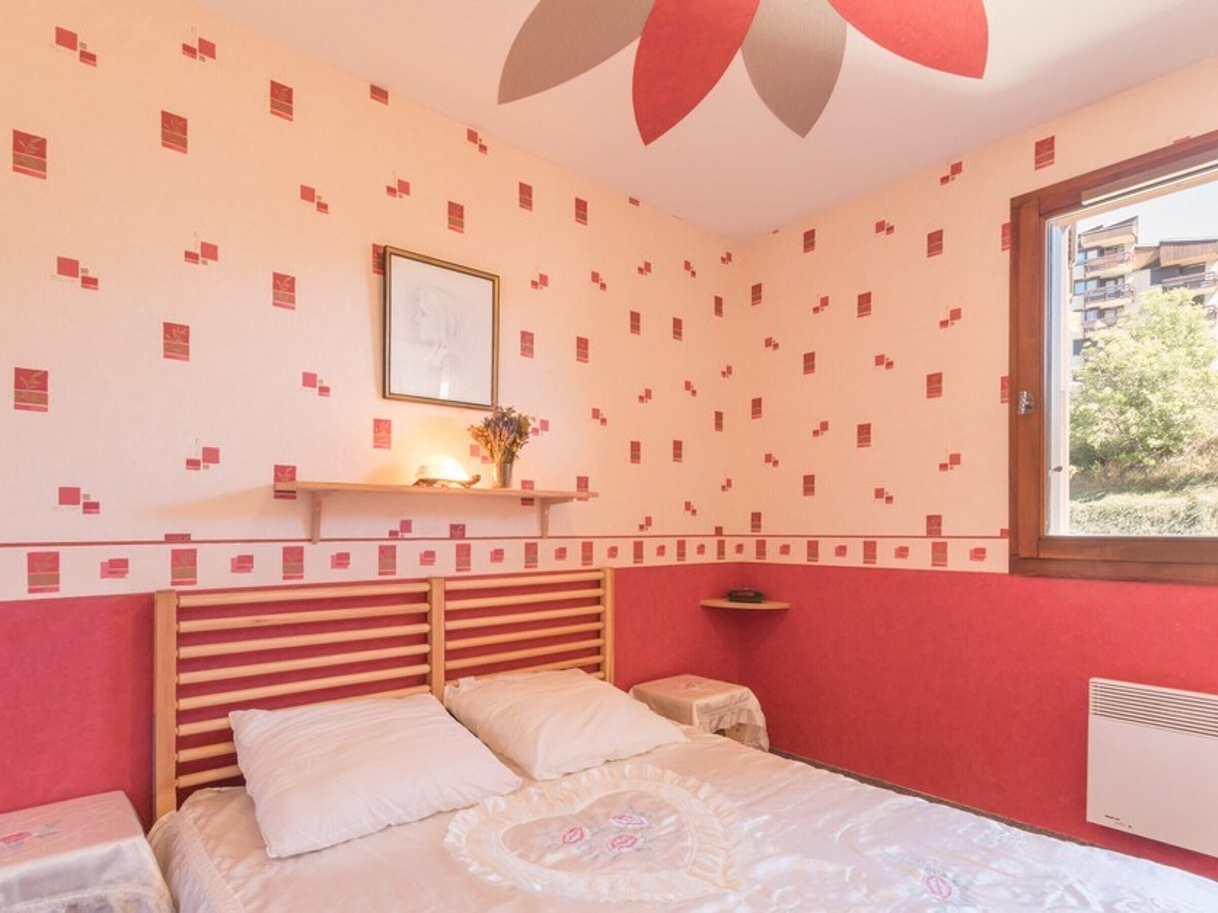 Une chambre à coucher comprenant un lit, une table de chevet, une étagère avec un tableau encadré et une fenêtre donnant sur des bâtiments et de la verdure.
