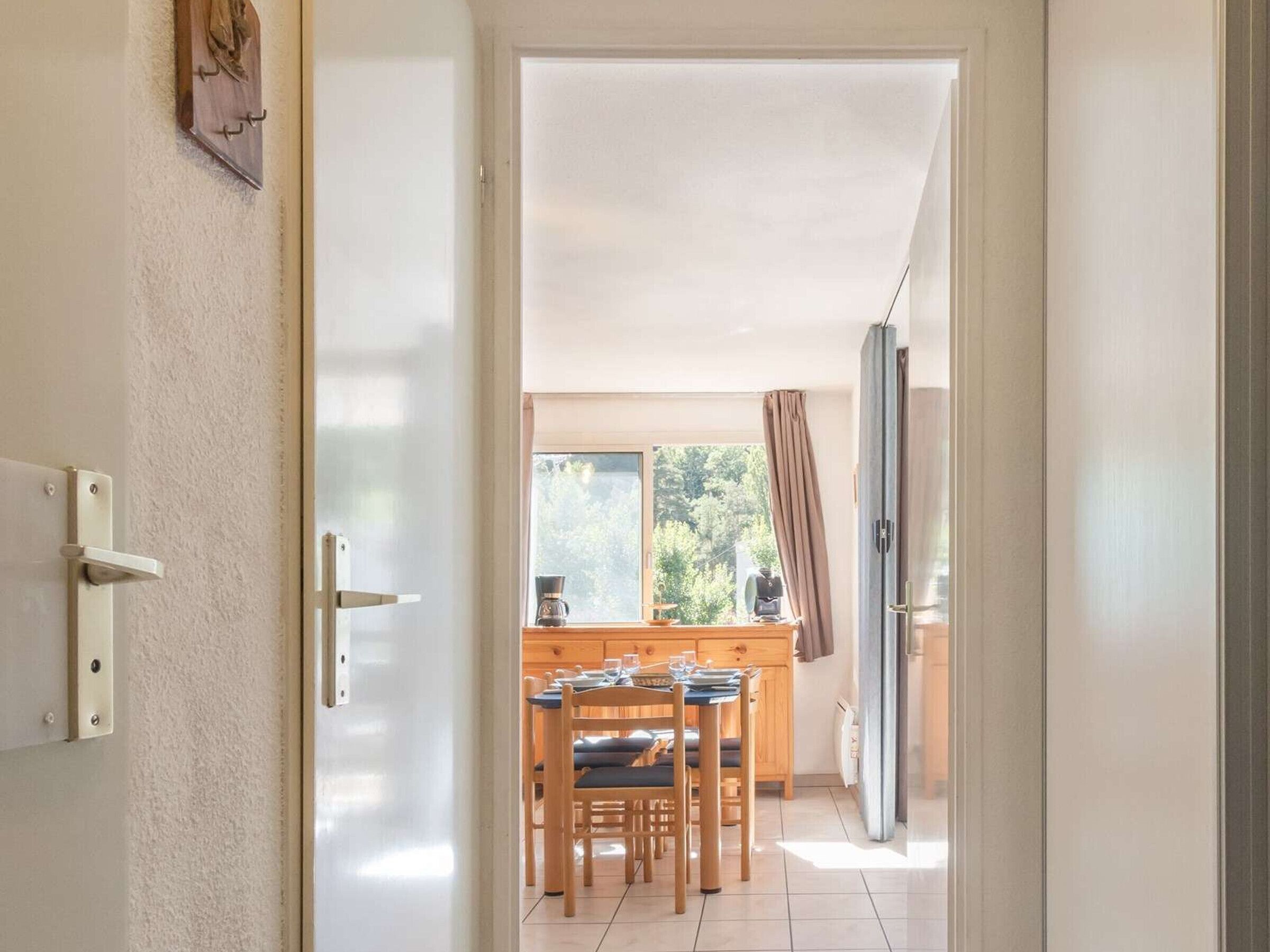 Une porte donne sur un coin repas, où se trouvent une table et des chaises en bois, une machine à café, et d’où l’on peut admirer les arbres à l’extérieur.