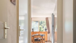 Un portal que dona a una zona de menjador amb una taula i cadires de fusta, una cafetera i vista a arbres a l'exterior.