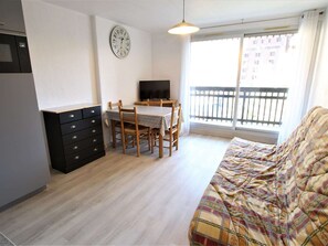 Um quarto pequeno e bem iluminado, com uma mesa de jantar e cadeiras, um sofá, uma televisão, um relógio e uma janela com vista para uma varanda.